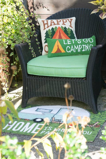 Chair with 'Happy Campers' pillows on a mat with 'Home is Where You Park It' text, surrounded by plants.