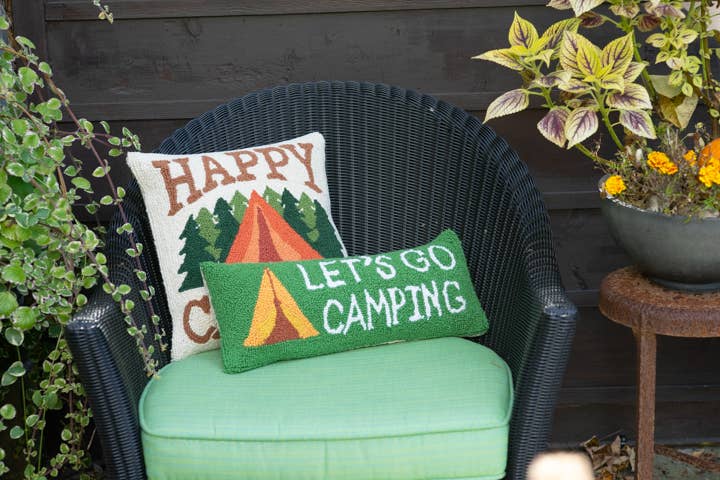 Green cushion with 'Let's Go Camping' text on a wicker chair outdoors.