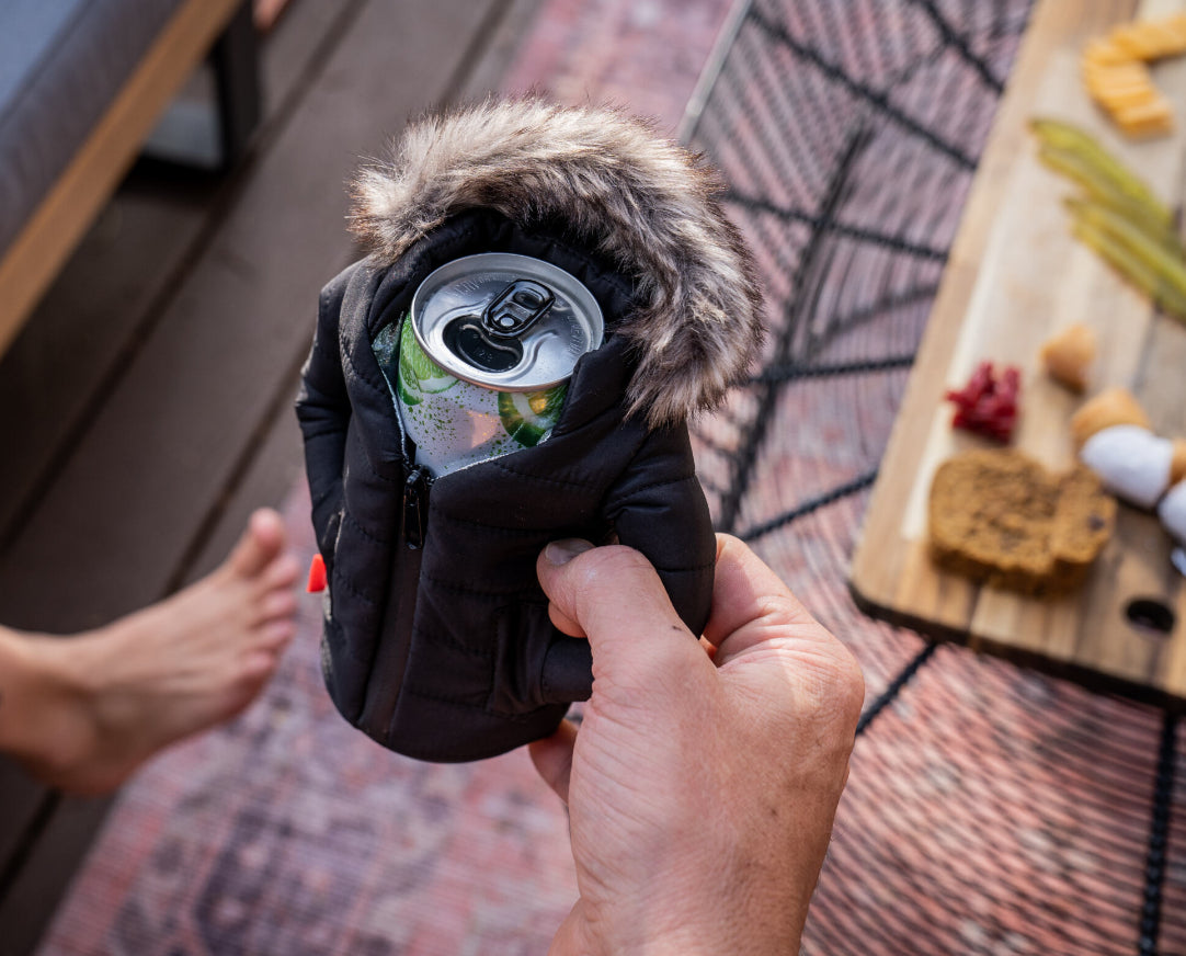 Hand holding a can of beer wrapped in a black jacket with fur trim on a wooden deck.