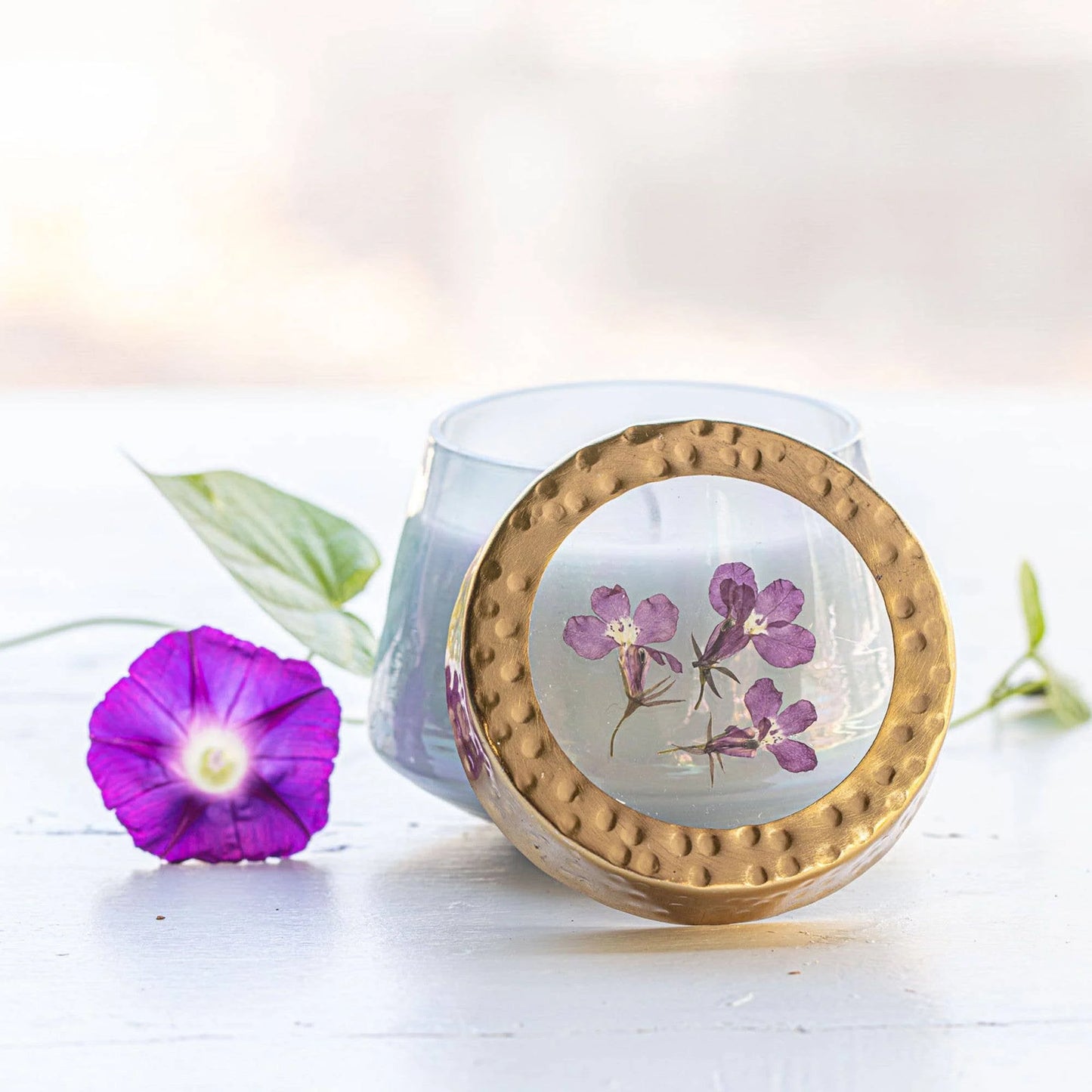 Candle with gold lid featuring pressed flowers, accompanied by a purple flower and green leaves on a light background.