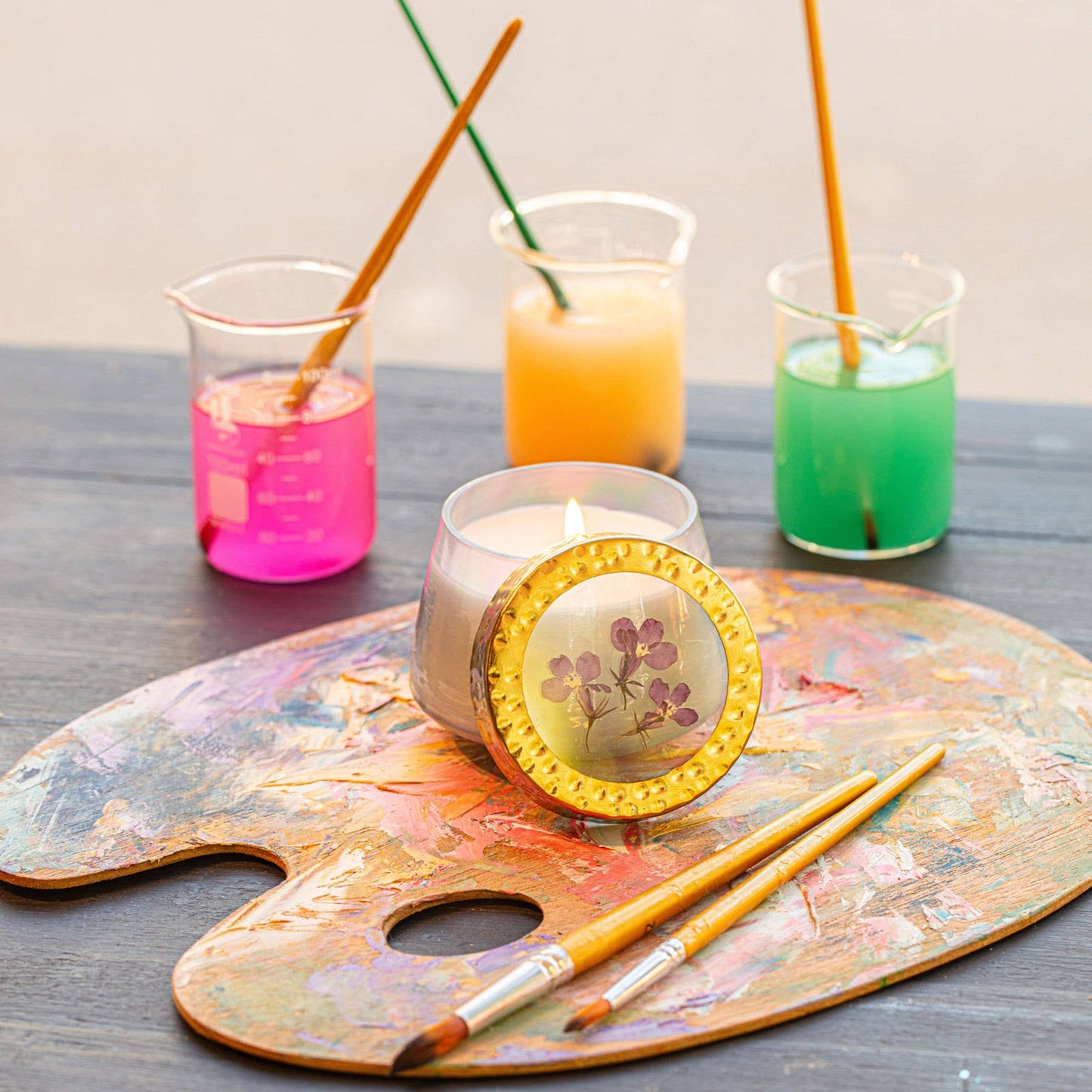Candle with a decorative cover on a paint palette with colorful liquids in beakers in the background.