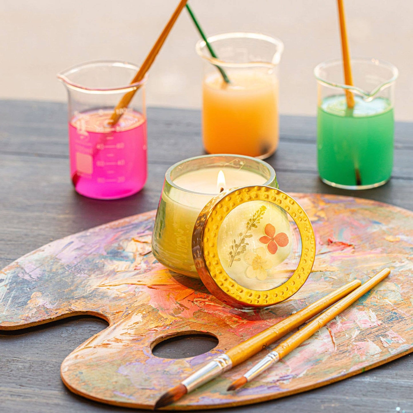 Candle with decorative cover on a paint palette with colorful drinks in the background