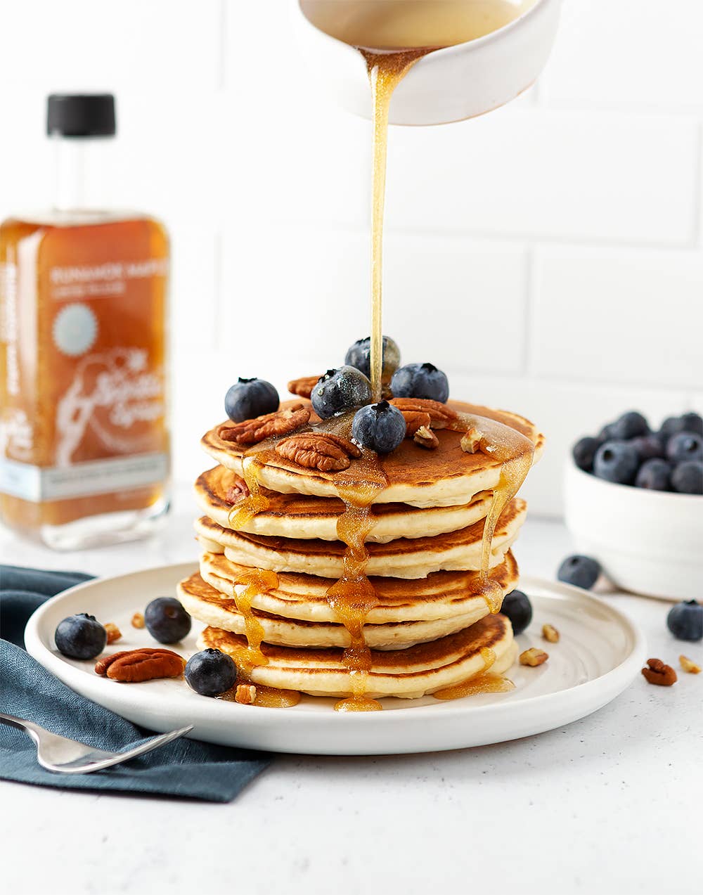 Stack of pancakes with syrup, blueberries, and pecans on a white plate with a bottle of syrup in the background.