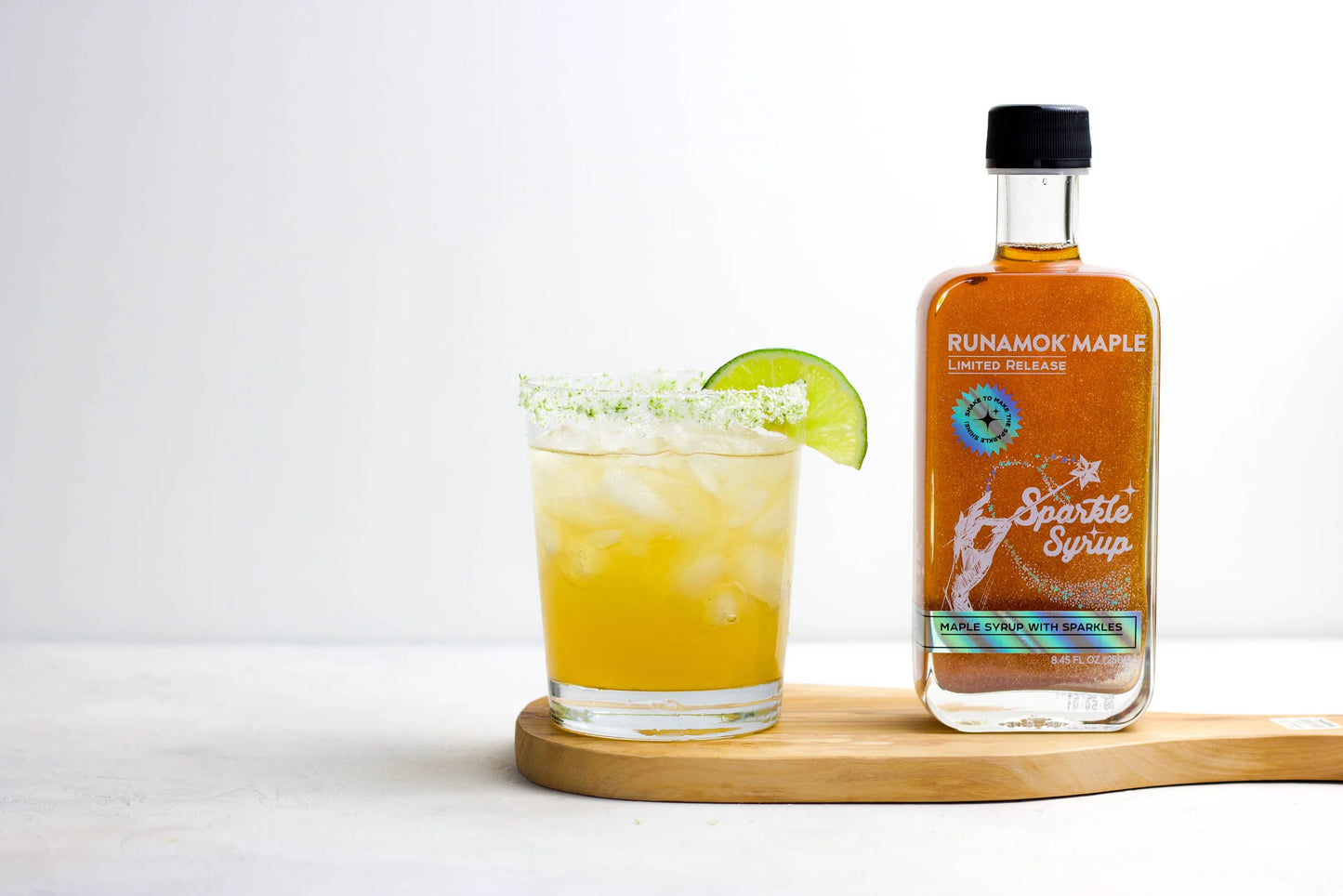 Bottle of Runamok Maple syrup next to a glass with a lime wedge on a white background