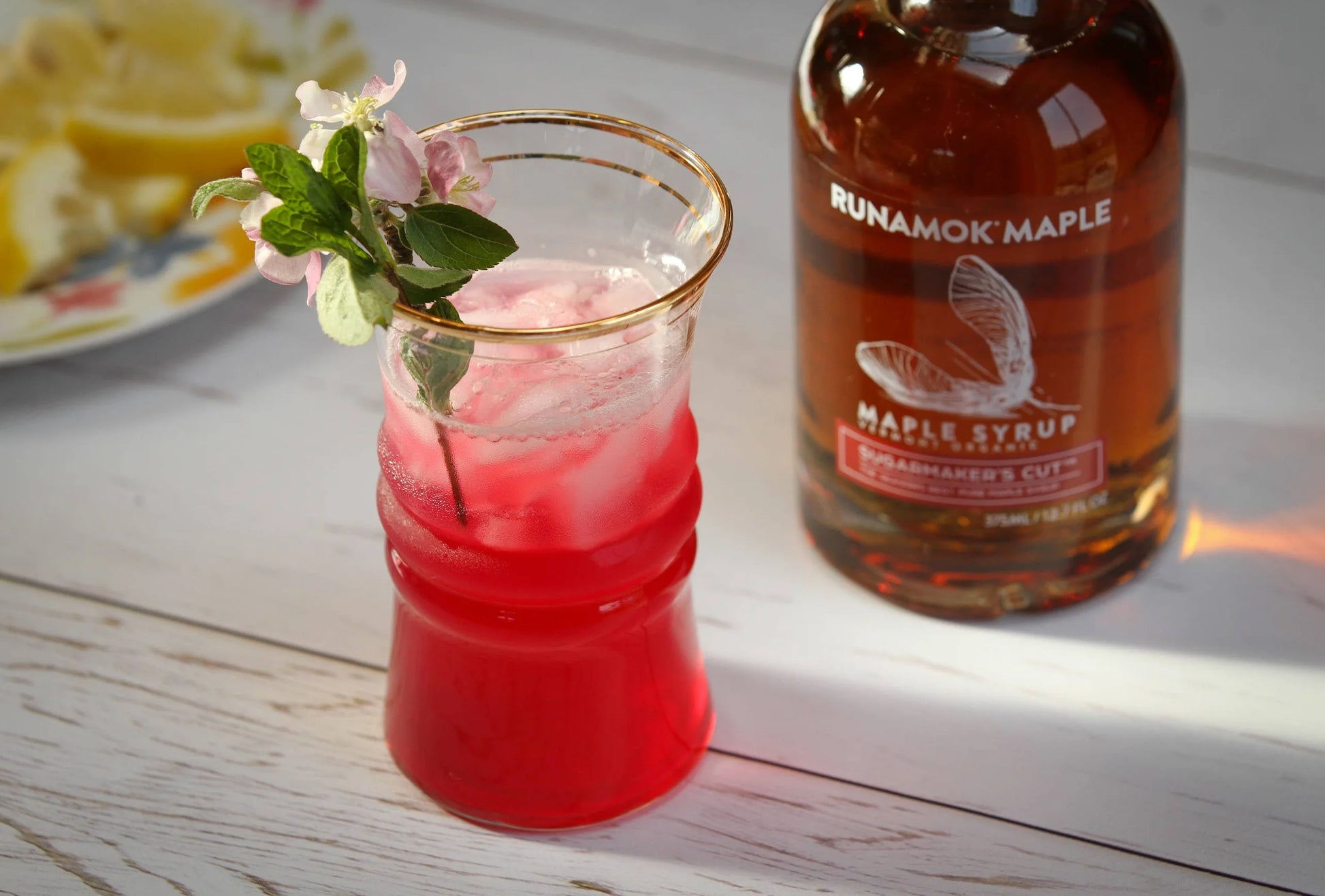 Red cocktail in a glass with ice and a sprig of mint next to a bottle of Runamok Maple syrup on a wooden surface.