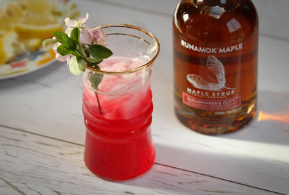 Red cocktail in a glass with ice and a sprig of mint next to a bottle of Runamok Maple syrup on a wooden surface.