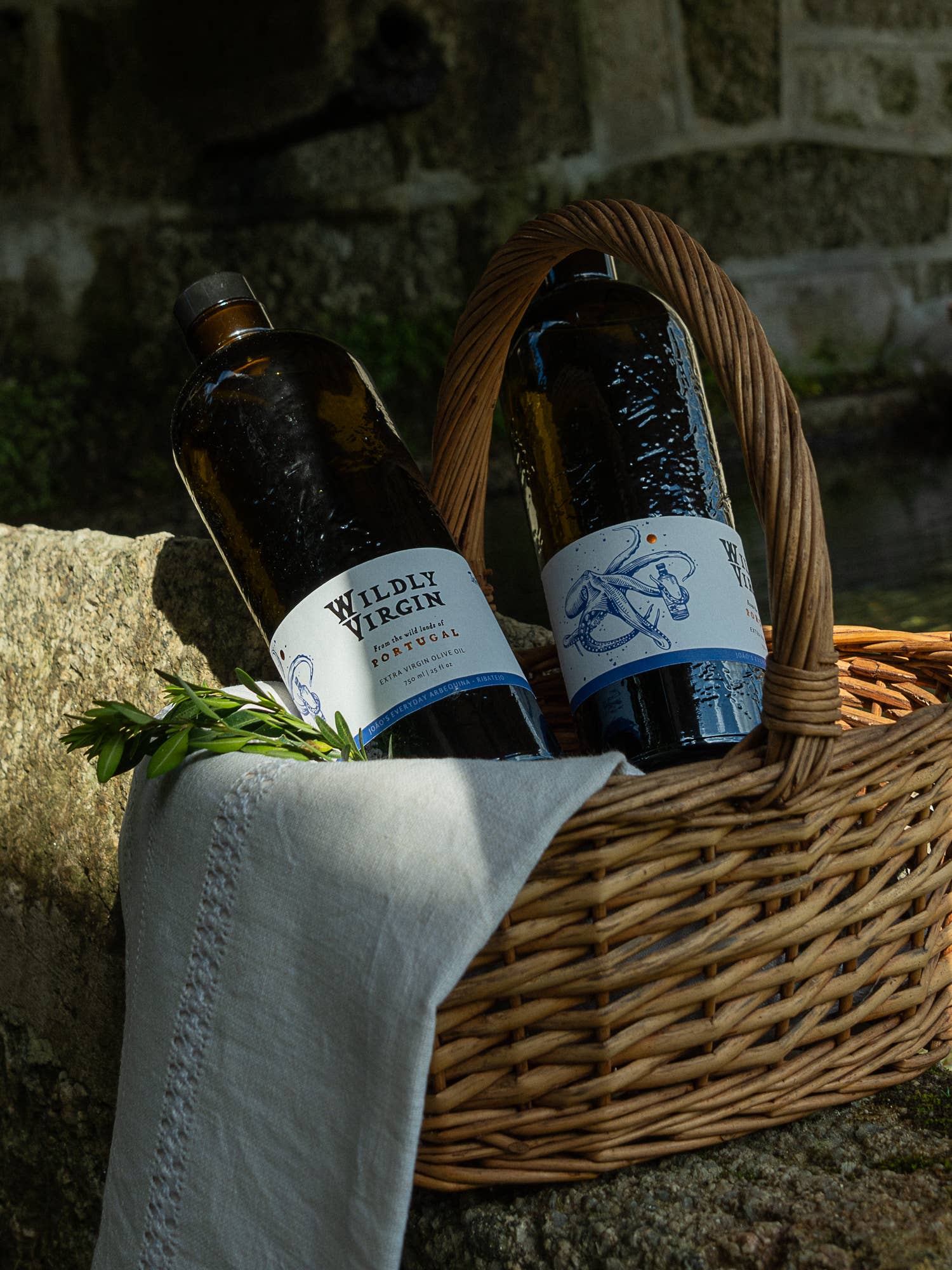 Two bottles of Wildly Virgin olive oil in a wicker basket with a stone wall background.