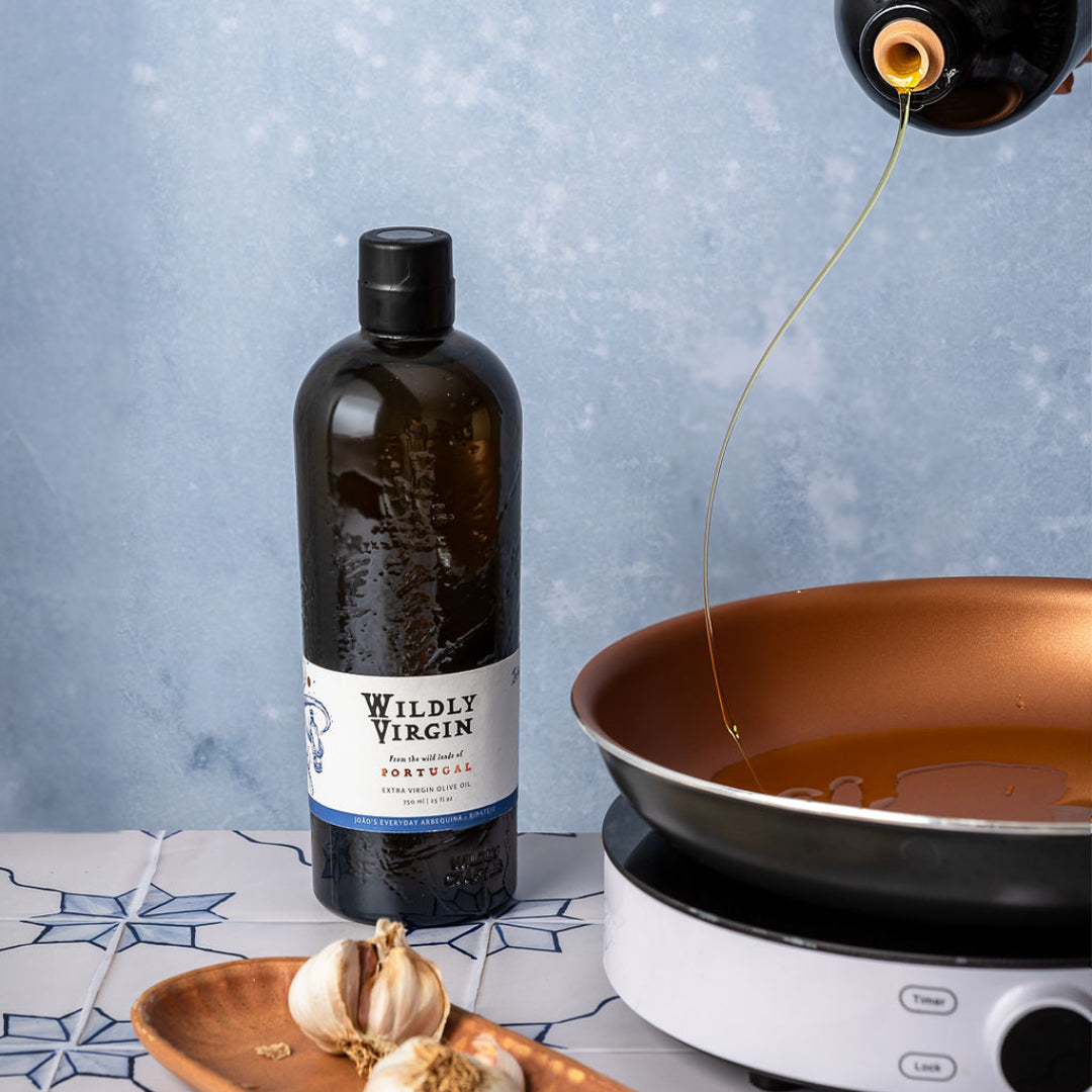Bottle of Wildly Virgin olive oil being poured into a pan with garlic on a tiled surface.