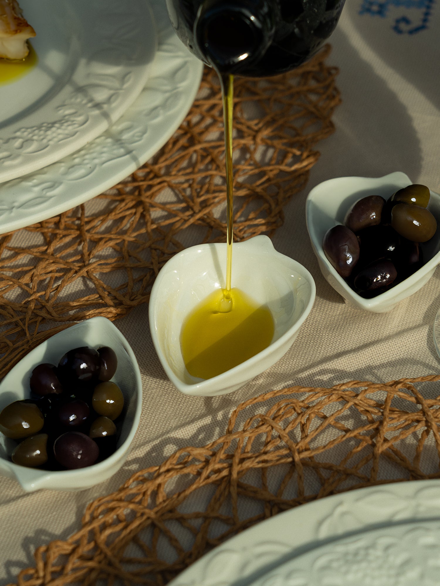 Olive oil being drizzled into a small white bowl with olives on a textured surface.