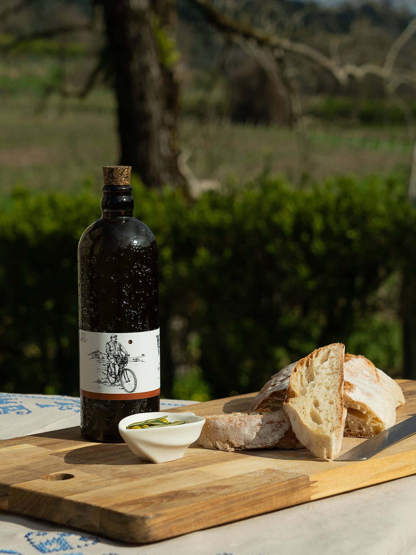 Bottle of olive oil with bread and a small dish on a wooden board outdoors.