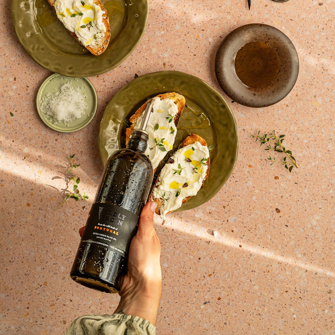 Person holding a bottle of olive oil next to a plate of bread with olive oil spread on a textured surface