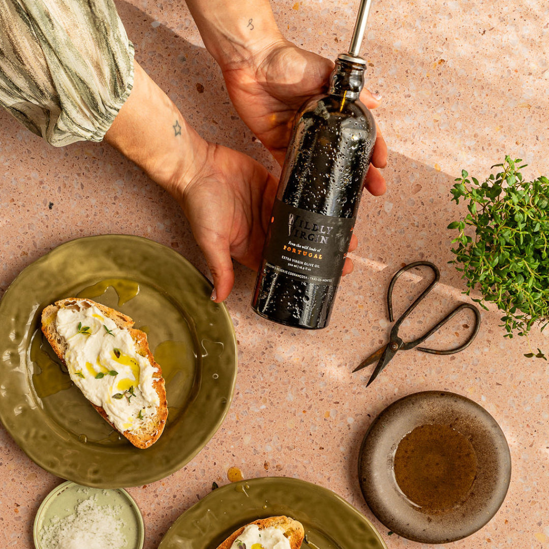 Person pouring olive oil from a bottle onto a plate with bread and herbs on a textured surface.